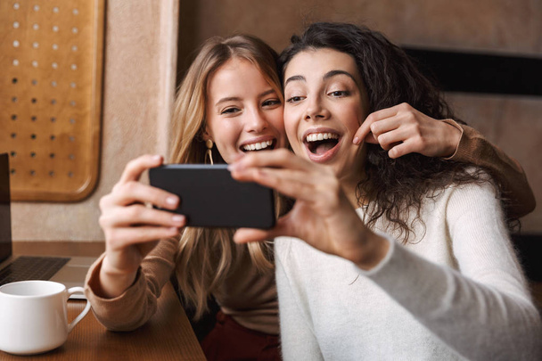 Excited happy pretty girls friends sitting in cafe using mobile phone. - Φωτογραφία, εικόνα