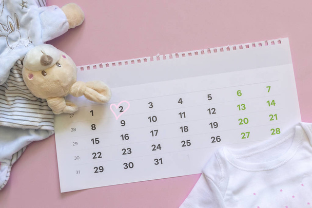 Set neugeborener Accessoires in Erwartung des Kindes - Kalender mit eingekreister Nummer 2 (zwei) Babykleidung, Spielzeug auf rosa Hintergrund. Kopierraum, flache Lage, Draufsicht. - Foto, Bild