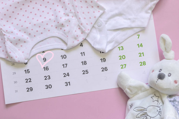 Комплект новорожденных аксессуаров в преддверии ребенка - календарь с кружком номер 9 (девять), детская одежда, игрушки на розовом фоне. Копировальное пространство, плоская кладка, вид сверху
. - Фото, изображение