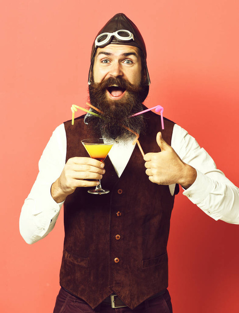handsome bearded pilot or aviator man with long beard and mustache on happy face holding glass of alcoholic beverage in vintage suede leather waistcoat with hat and glasses on red studio background - Fotoğraf, Görsel