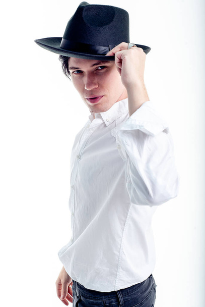Young brunette man in white shirt and black hat posing on white background  - Фото, изображение