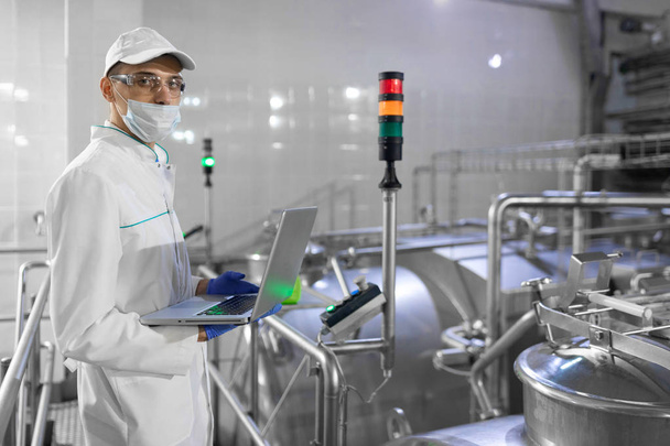Retrato del hombre con una bata blanca y una gorra de pie en el departamento de producción de la fábrica de lácteos con ordenador portátil
 - Foto, imagen