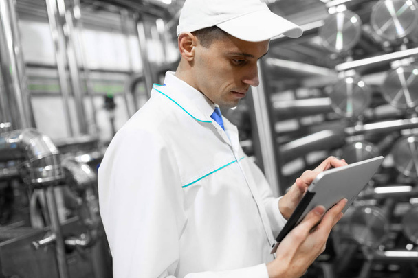 Portret van de mens in een wit gewaad en een pet staande in productieafdeling van zuivelfabriek met grijze Tablet - Foto, afbeelding