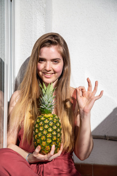 Nuori iloinen aistillinen nainen rento kesä mekko tilalla trooppinen ananas suljetuin silmin ja osoittaa ok ele
 - Valokuva, kuva