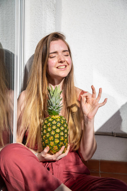 Young cheerful sensual woman in casual summer dress holding tropical pineapple with closed eyes and showing ok gesture - Фото, зображення