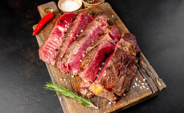 Čerstvé vzácné grilované bifteky s kořením na dřevěné sekací desce přes černé kamenné pozadí - Fotografie, Obrázek