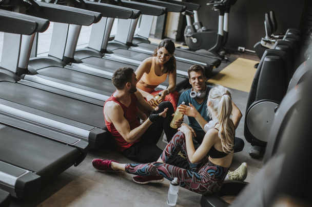 Grupa młodych ludzi w odzieży sportowej rozmawiając i śmiech razem siedząc na podłodze siłowni po treningu - Zdjęcie, obraz