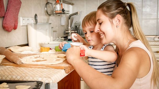 Retrato de primer plano del adorable niño de 3 años haciendo galletas con madre. CHild sosteniendo la bandeja para hornear y poniendo galletas en ella
 - Foto, imagen