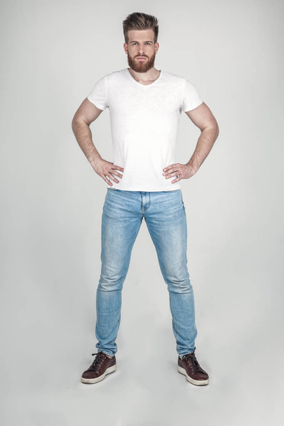 A handsome man with a beard and a beautiful hairstyle dressed in casual clothing looks at the camera. stands opposite the white background - Photo, Image