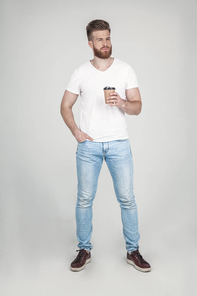 A beautiful man with a beard and a beautiful hairstyle, dressed in casual clothes, holds a cup of coffee and looks away stands opposite the white background - Foto, afbeelding