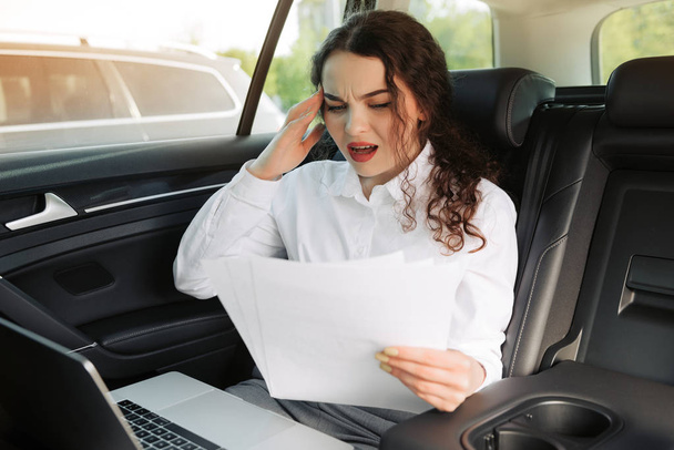 Verwirrte Geschäftsfrau liest Hiobsbotschaft und arbeitet im Auto mit Dokumenten und Laptop. - Foto, Bild