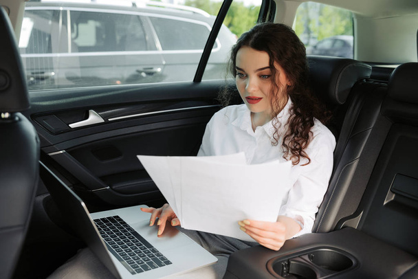 Όμορφη νεαρή επιχείρηση γυναίκα χρησιμοποιώντας φορητό υπολογιστή και δουλεύοντας με τα έγγραφα στο αυτοκίνητο. - Φωτογραφία, εικόνα