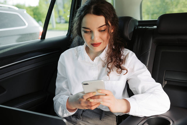 Όμορφη νεαρή επιχείρηση γυναίκα χρησιμοποιώντας το laptop και το τηλέφωνο στο αυτοκίνητο. - Φωτογραφία, εικόνα