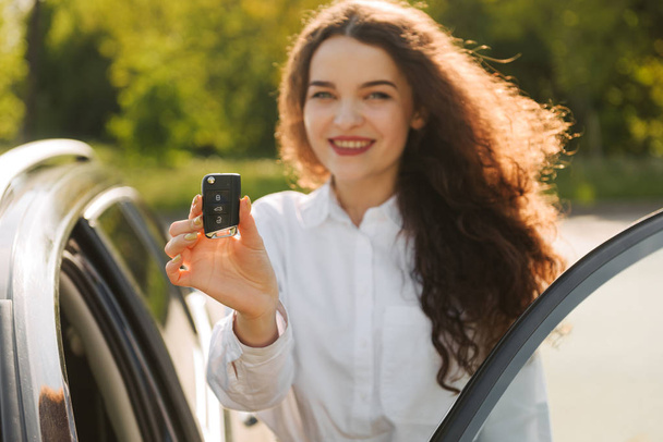 Outdoor shot of satisfied beautiful female owner of car, likes and rejoices her purchase, holds keys , has happy expression, stands near new automobile, going to drive - Fotó, kép