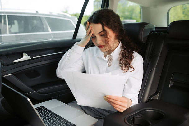 Расстроенная деловая женщина, читающая плохие новости, работающая с документами и ноутбуком в машине
. - Фото, изображение