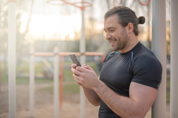 handsome male athlete, in the summer outside on the sports ground, smiling, typing sms, using a smartphone - Photo, Image