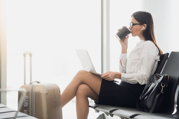 Young businesswoman in the airport, using laptop and drinking coffee, travel, business trip and active lifestyle concept - Photo, Image