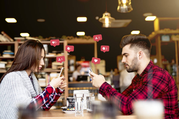 Привлекательная кавказская пара одета непринужденно, используя смартфоны для социальных сетей, сидя в кафе
. - Фото, изображение
