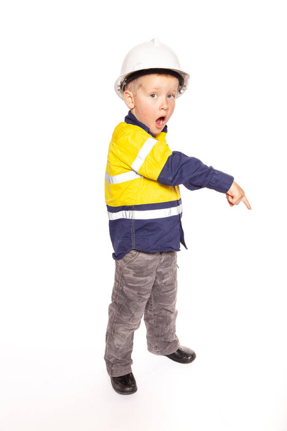 Young blond caucasian boy pointing down with a shocked look role playing as a construction worker supervisor in a yellow and blue hi-viz shirt, boots, without the white hard hat, a hammer and tape measure. - Zdjęcie, obraz