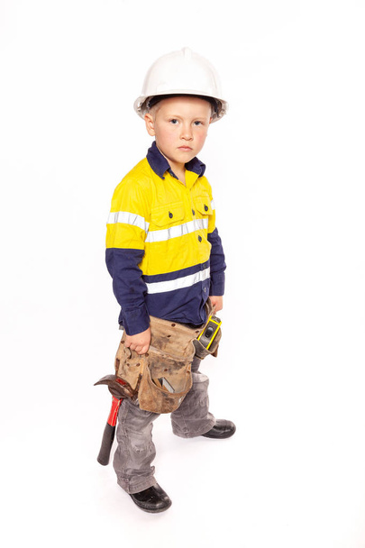 Young blond caucasian boy looking out with a fierce look role playing as a construction worker supervisor in a yellow and blue hi-viz shirt, boots, white hard hat, tool belt, a hammer and tape measure. - 写真・画像