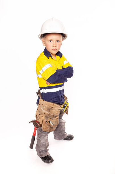Young blond caucasian boy arms folded or on his hips role playing as an angry construction worker in a yellow and blue hi-viz shirt, boots, white hard hat, tool belt, a hammer and tape measure. - Foto, Imagen