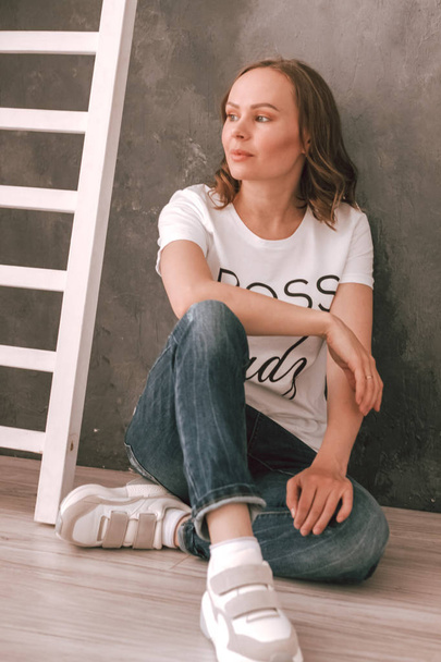 Photo of a young woman in a white t-shirt, jeans against a dark wall sitting on the floor - Photo, Image