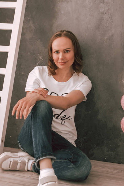 Photo of a young woman in a white t-shirt, jeans against a dark wall sitting on the floor - Фото, изображение