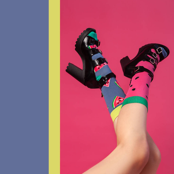 Konzept der fröhlichen Sommerstimmung, Entspannung und Schönheit. Frauenbeine in schwarzen Schuhen mit Absätzen und hellen Socken mit Wassermelonenaufdruck und Samen auf einem isolierten leuchtend rosa, gelben, blauen Hintergrund - Foto, Bild