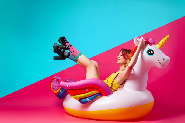 Konzept Sommerstimmung, Entspannung und Schönheit. Modernes Porträt einer jungen Frau im gelben Badeanzug, hellen Socken und Sandalen, die auf einer aufblasbaren Einhornmatratze auf einem isolierten rosa-blauen Hintergrund ruhen. - Foto, Bild