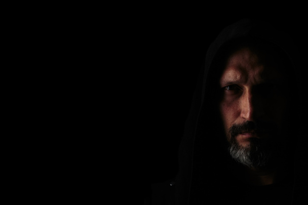 Brutal man with a gray beard in the hood with sharp shadows on a black background. Copy space. - Photo, Image