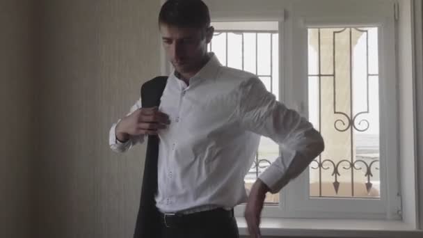 man with dark hair puts on waistcoat on shirt against window - Záběry, video