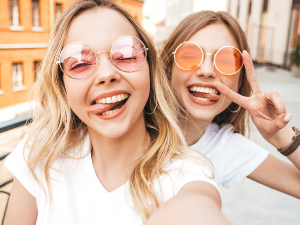 Two young smiling hipster blond women in summer white t-shirt clothes. Girls taking selfie self portrait photos on smartphone.Models posing on street background.Female shows peace sign and tongue - Photo, image