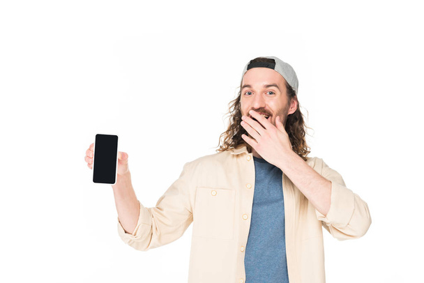 excited young man holding smartphone and closing mouth with hand isolated on white - Foto, imagen