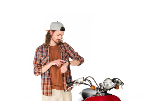young man looking and pointing with finger at watch while standing near red scooter isolated on white - Zdjęcie, obraz