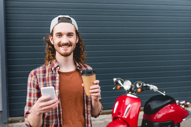 joven sosteniendo taza de papel con café y teléfono inteligente, de pie cerca de scooter rojo
  - Foto, Imagen