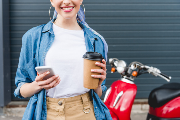 vista recortada de la hermosa chica de pie cerca de scooter rojo, sosteniendo taza de papel con café y teléfono inteligente
 - Foto, Imagen