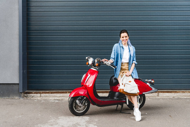 full length view of beautiful girl sitting on red scooter and holding backpack - Foto, imagen
