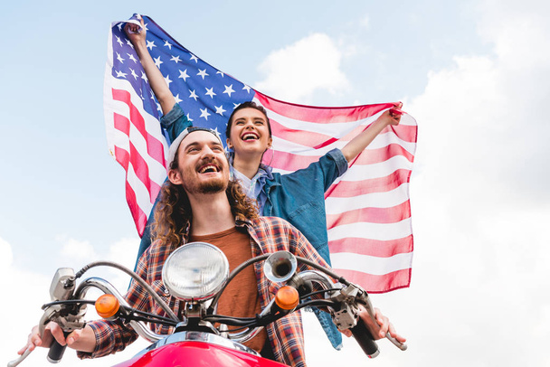lage hoek weergave van mooi meisje rijden met jonge man op rode scooter en holding Amerikaanse vlag - Foto, afbeelding