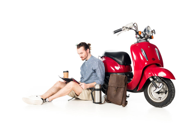  young man holding paper cup, looking at map and sitting near red scooter isolated on white - 写真・画像