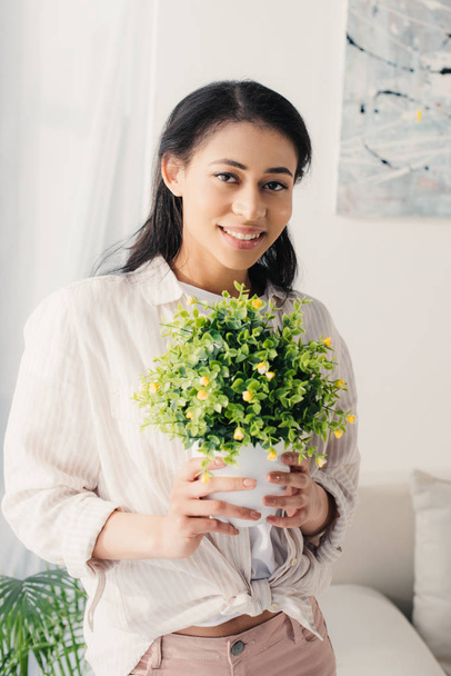 pretty latin woman holding flowerpot with green plant while smiling at camera - Фото, зображення