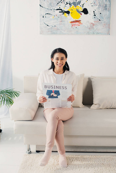 hermosa mujer latina sosteniendo periódico de negocios mientras está sentado en el sofá y sonriendo a la cámara
 - Foto, imagen