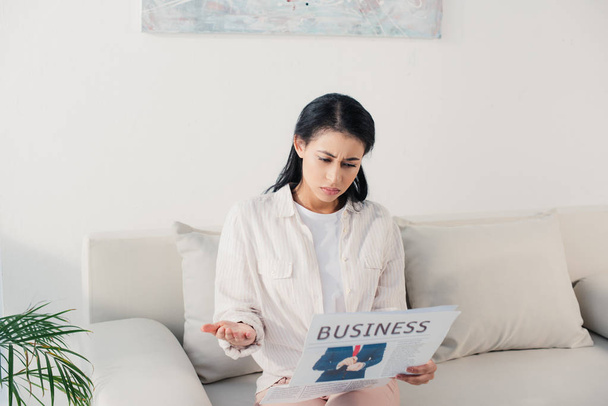 shocked latin woman reading business newspaper while sitting on sofa at home - Foto, Bild
