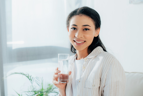 positive latin woman holding glass of water while smiling at camera - Photo, Image