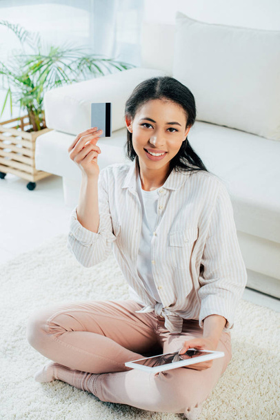 aantrekkelijke jonge vrouw met behulp van digitale Tablet en het houden van creditcard terwijl zittend op de vloer en glimlachend op de camera - Foto, afbeelding