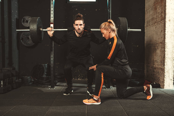 νεαρός μυώδης άνδρας κάνει ασκήσεις squat με barbell με προσωπικό γυμναστή στο γυμναστήριο - Φωτογραφία, εικόνα