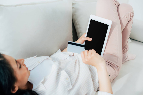 young latin woman holding credit card and digital tablet with blank screen while lying on sofa at home - Fotografie, Obrázek