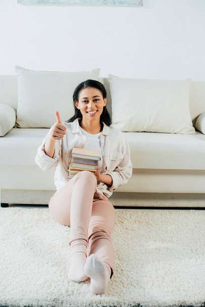 cheerful latin woman showing thumb up and holding stack of books while sitting on floor at home - Photo, Image