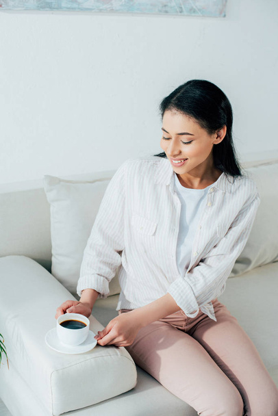 cheerful latin woman holding cup of coffee while sitting on couch at home - Fotó, kép