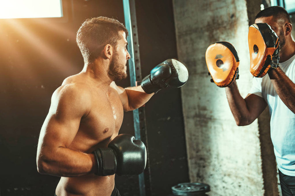 Νεαρός μυώδης άνδρας που κάνει ασκήσεις πυγμαχίας με προσωπικό γυμναστή στο γυμναστήριο - Φωτογραφία, εικόνα