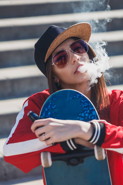 Vaping girl. Young woman with skateboard vape e-cig. Pretty young female in black hat, red clothing vape ecig, vaping device at the sunset. Toned image. Hip-hop style. - Photo, Image